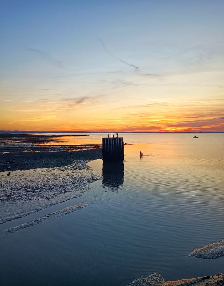 A photo of the sea and sky at dusk. The sun has set and the sky is orange near the horizon. In the centre of the vertical image, a dark stack-like structure rises. A man with a large net is walking in the water close by. The sky is reflected in the water's surface.