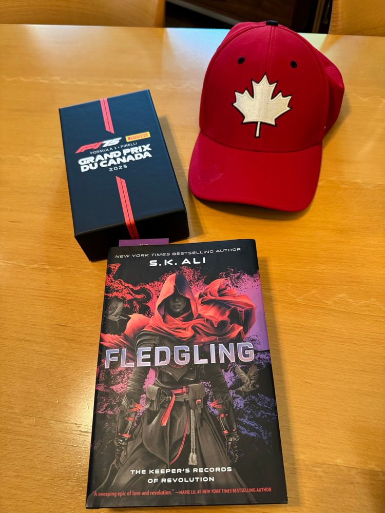 A black box labeled Formula 1 Grand Prix Du Canada, a red baseball cap with a Canadian flag, a hardcover book called Fledgling by S.K, Ali