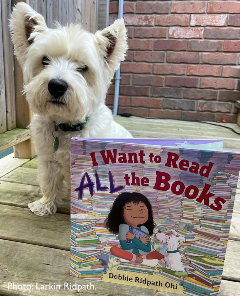 A fluffy white West Highland Terrier (Westie) sits on a wooden deck next to a children's book titled I Want to Read ALL the Books by Debbie Ridpath Ohi. The book cover features an illustration of a child happily holding a book, surrounded by stacks of books, with a white dog beside them. The scene suggests a love for reading and is warm and inviting, enhanced by the cute dog in the photo. Photo credit: Larkin Ridpath.