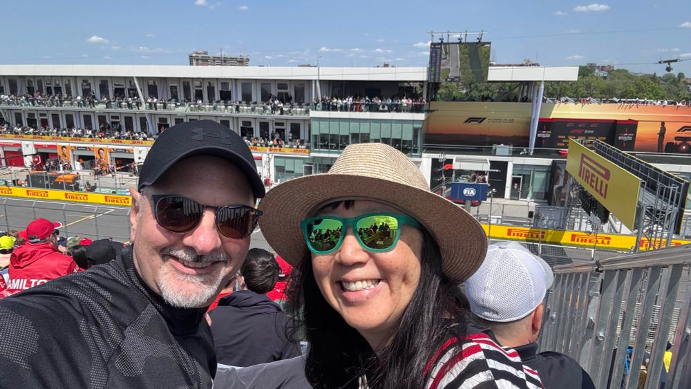 A smiling couple takes a selfie in the grandstands at a Formula 1 event. The track, pit lane, and Pirelli-branded barriers are visible behind them, along with team garages, a crowd-filled building, and a large digital screen showing race footage and standings. The weather is sunny with a clear blue sky.