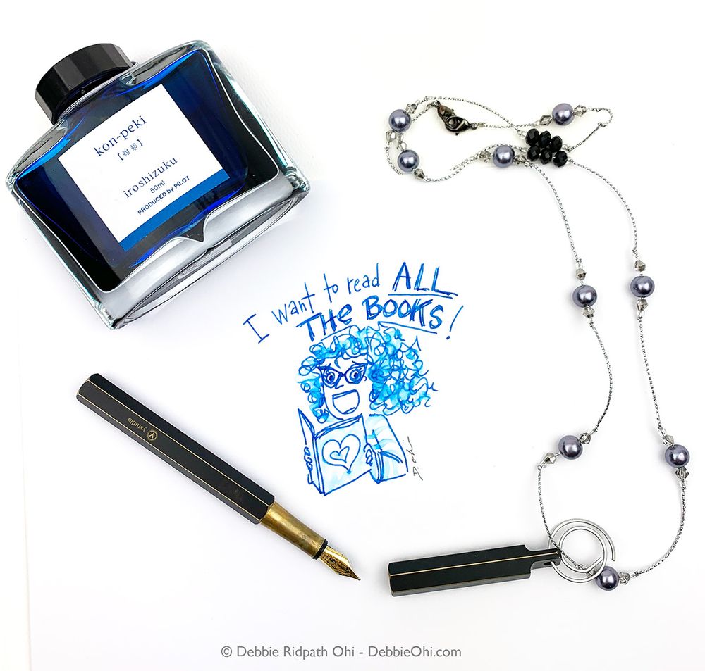 Overhead photo showing a bottle of Pilot Iroshizuku Kon-peki blue ink, a black and gold YStudio fountain pen with the cap removed, and a silver necklace with dark pearls. In the center is a blue ink drawing of an excited curly-haired girl holding a book, with the caption “I want to read ALL THE BOOKS!” written above. Illustration is signed by Debbie Ridpath Ohi.
