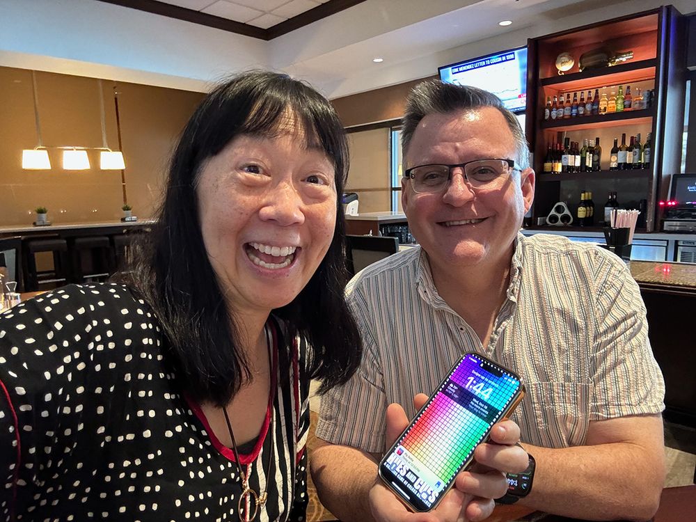 Two smiling individuals seated at a bar, with one holding a phone whose lock screen includes a colorful grid pattern. The background features a stocked bar, pendant lights, and a TV screen.