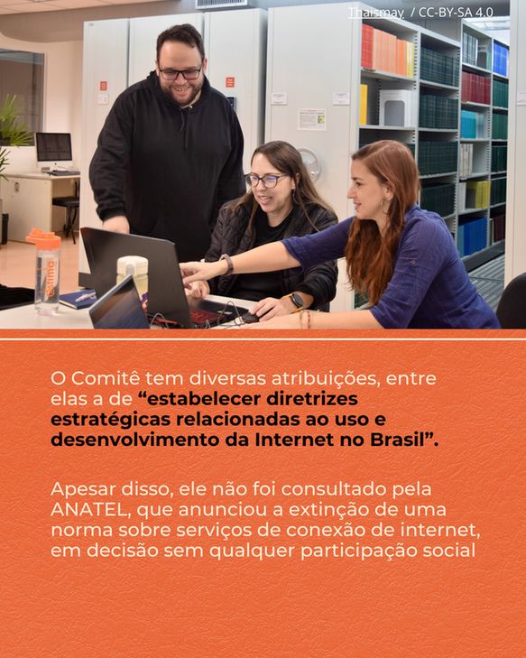 Fotografia de três pessoas em um ambiente que parece ser um escritório ou biblioteca, com estantes de arquivos ao fundo. Um homem de óculos e barba, vestindo uma blusa escura, está em pé atrás de uma mesa, olhando para um laptop e sorrindo. Sentadas à sua frente, duas mulheres interagem com o laptop; uma, de óculos e blusa escura, olha para a tela, enquanto a outra, de blusa azul, aponta para o computador. Sobre a mesa, há também outro laptop menor e material de escritório. A metade inferior da imagem é um bloco de cor laranja com texto em branco. O primeiro parágrafo diz: "O Comitê tem diversas atribuições, entre elas a de “estabelecer diretrizes estratégicas relacionadas ao uso e desenvolvimento da Internet no Brasil”.". O segundo parágrafo afirma: "Apesar disso, ele não foi consultado pela ANATEL, que anunciou a extinção de uma norma sobre serviços de conexão de internet, em decisão sem qualquer participação social".