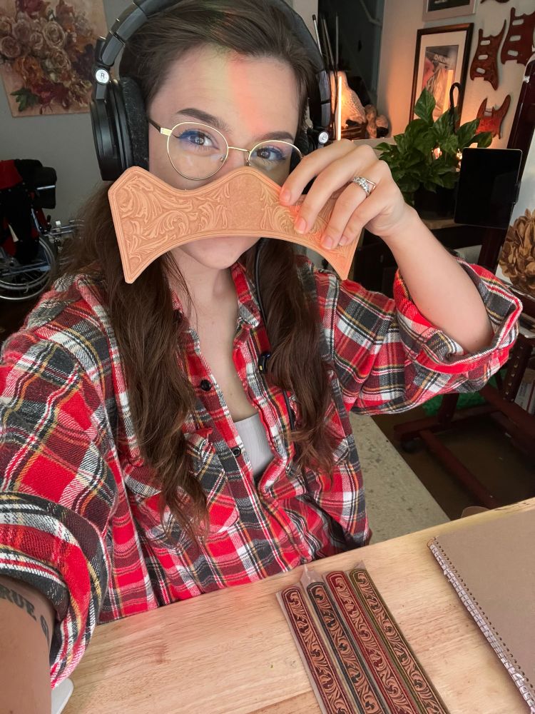 Selfie of me at my workbench wearing a plaid holding up a piece of tooled leather infront of my face