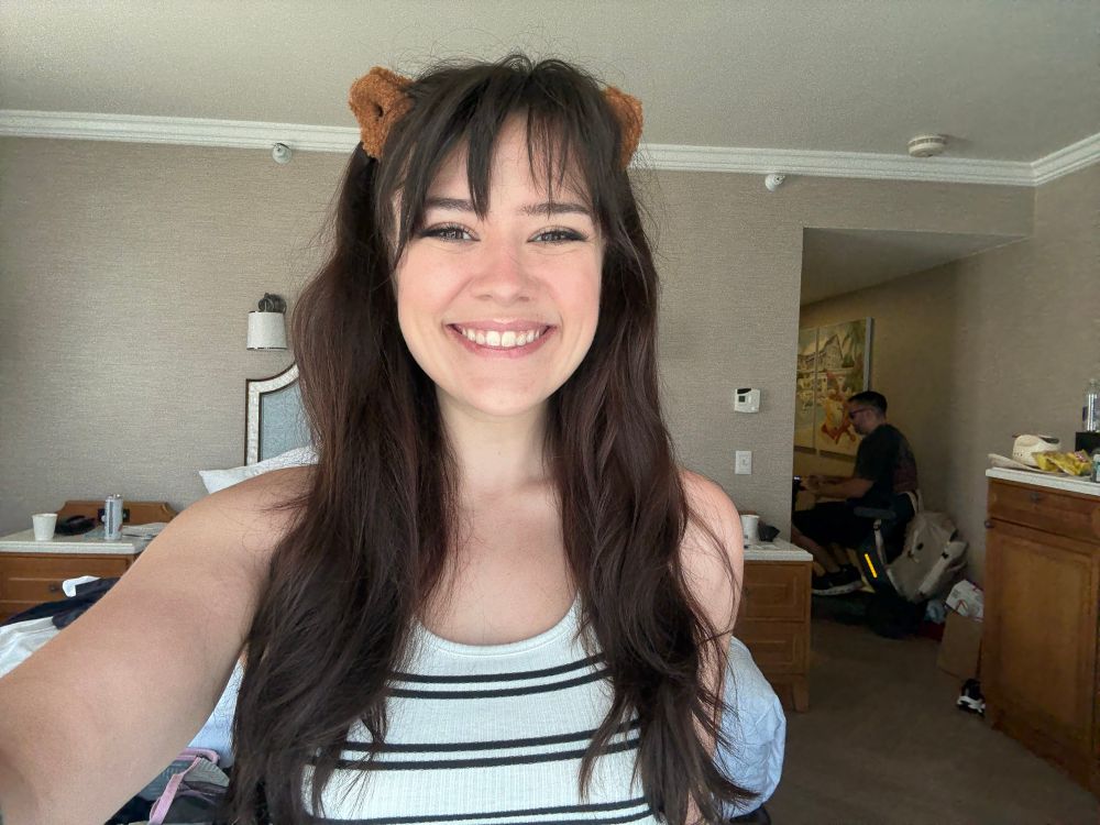 Selfie of me with long brown hair and bear ears with my husband in the background on my scooter