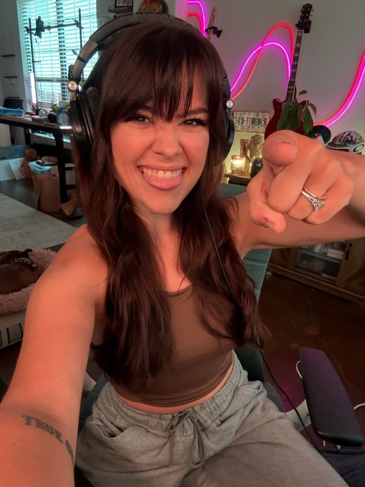 Selfie of em at my desk biting my tongue and pointing at the camera 
