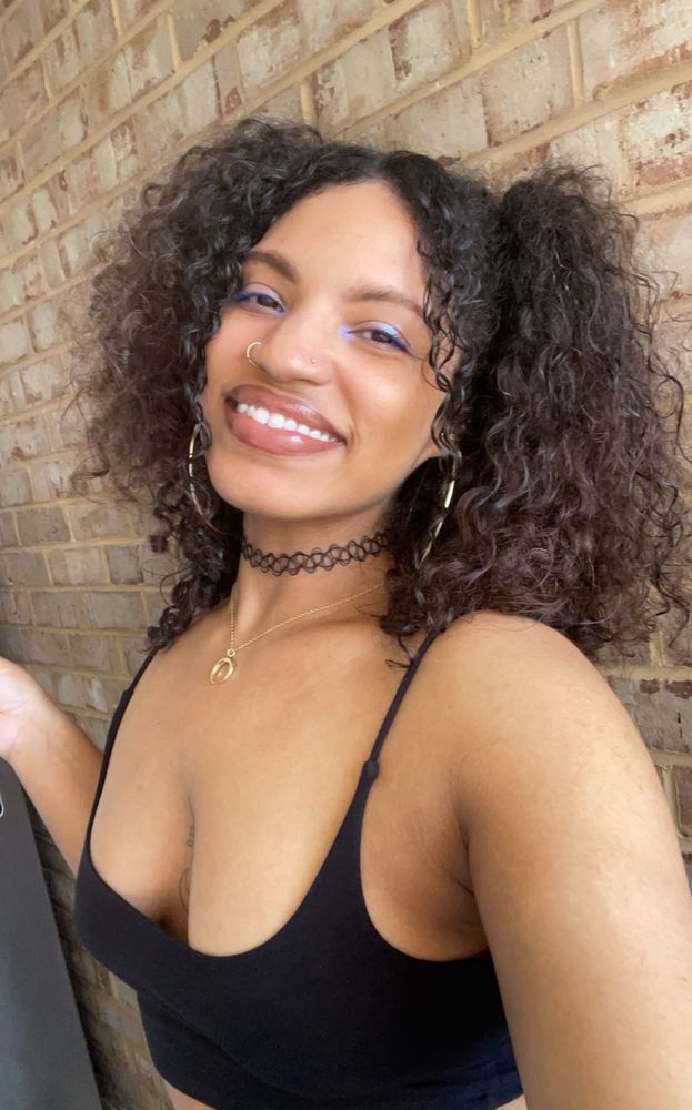 selfie smiling in front of a brick wall, black cropped tank, black choker, gold moon necklace, curly hair in pigtails half up half down, gold hoops, gold nose rings, and blue shimmer on the eyes with blue mascara