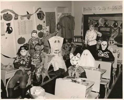 A black and white photo of a classroom from 1960s. The kids are costumed for Halloween. A cavalry corporal, a clown?, someone I can't identify, a kid holding a magazine against his face?, a fox, a ghost, a skeleton, a giant bunny, and the adult in the middle I think is the teacher.