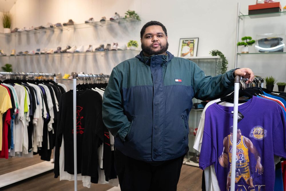 A man wearing a green and navy jacket stands in a clothing store with racks of shirts, including a purple Lakers shirt, and shoes displayed on shelves behind him.