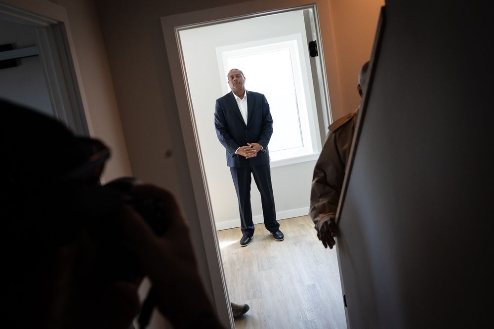 A man in a suit stands in a bright, empty room framed by a doorway, while silhouettes of people are visible in the foreground.