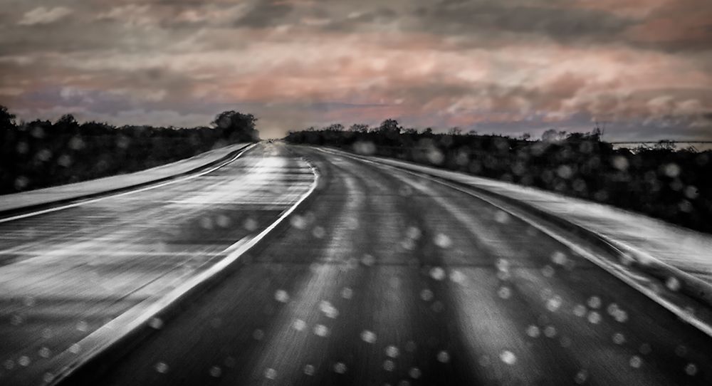 View of a rainy windshield and a lonely highway from inside a car with pink and gray pastel clouds on the horizon.