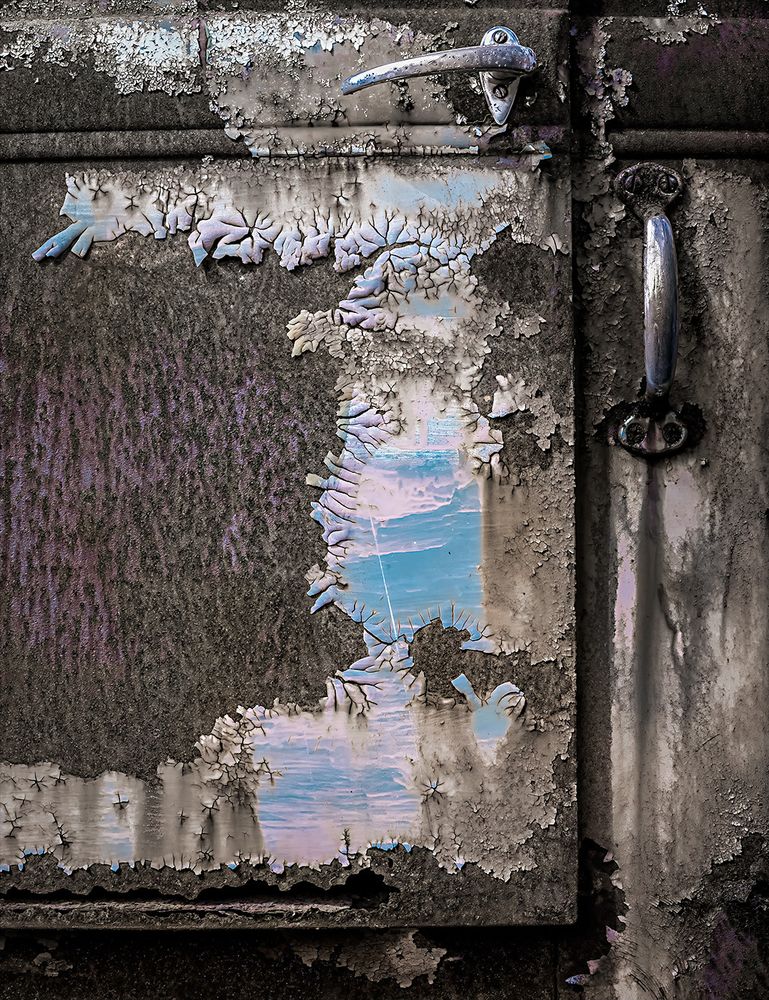 Blue and pink paint peeling on a rough and rusted truck door, bracketed by chrome door handles.