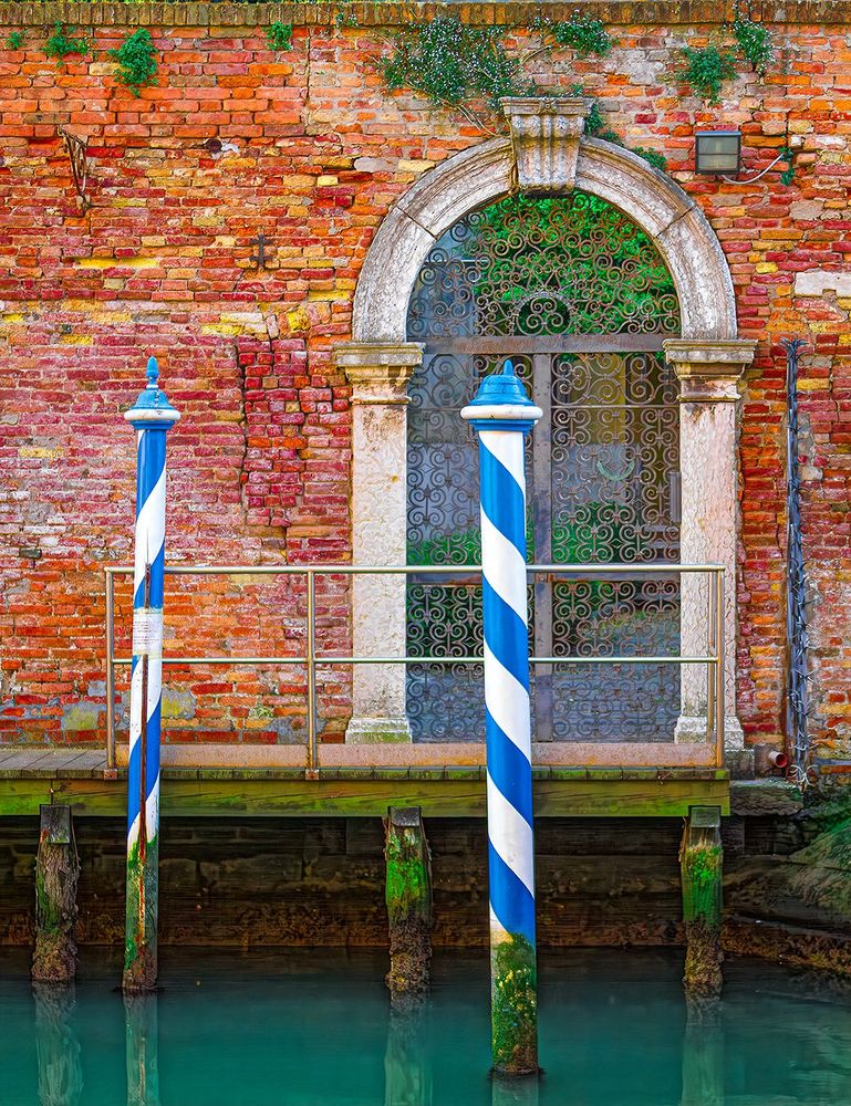 Arched doorway on brick building in Venice, Italy, leading to a dock on the canal with colorful tie-up posts.