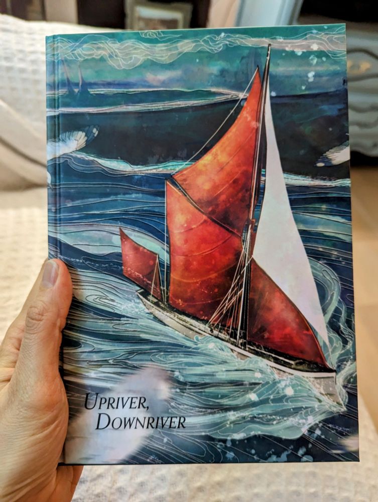 A hand holds the book of the new ttrpg, Upriver, Downriver. The cover is of a ship on water.
