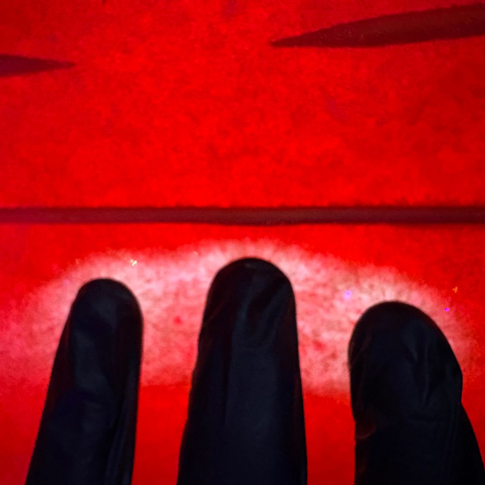 Three gloved fingers pressing down gently on a clear gel plate causing the red liquid underneath to whiten under the fingertips. Three wooden spikes jut into the picture above the fingers, one cutting the photo in half and two ends sticking in from either side. 