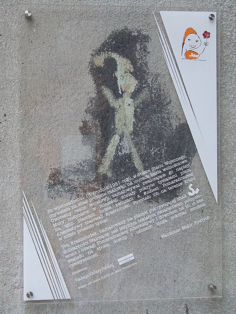 Orange Alternative Dwarf on Madalińskiego Street in Warsaw, preserved under an acrylic sheet printed with text in Polish outlining the use of gnomes as an absurdist means of resistance against martial law in Poland in the 1980s