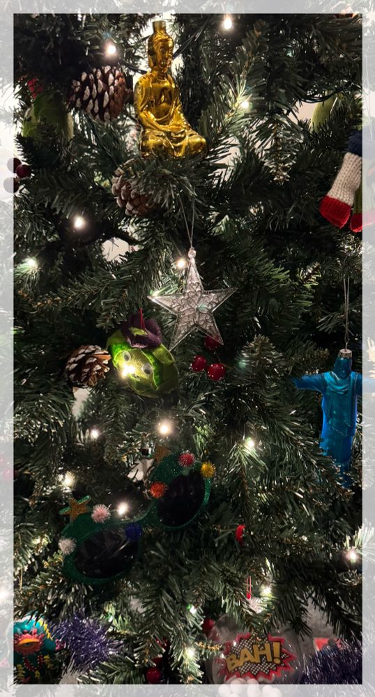 A rectangular photo of a portion of a Christmas tree, with a golden Buddha, Hamsa hand, blue kitsch Jesus, bah! cut out in a giant bauble and others, with twinkly lights. 