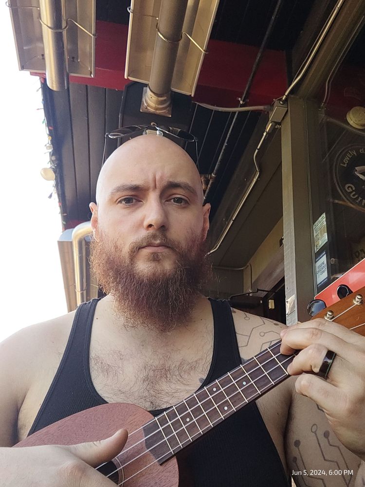Bald man with beard holding an ukulele at a pub. He is holding an E7 chord mid strum.