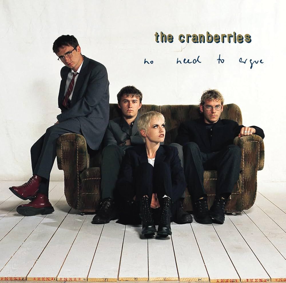 Couverture de l'album "No Need to Argue" des Cranberries.
On voit le groupe dans une piece entièrement blanche, assis sur un petit canapé marron.
La chanteuse est assise par terre devant, elle regarde vers le coin haut droit de l'image, et les autres membres du groupe nous regardentt.
La chanteuse Dolores O'Riordan est décédée en 2018 (il y a déjà 6 ans...). Une des plus belles voix de son époque et peut-être au-delà. 
