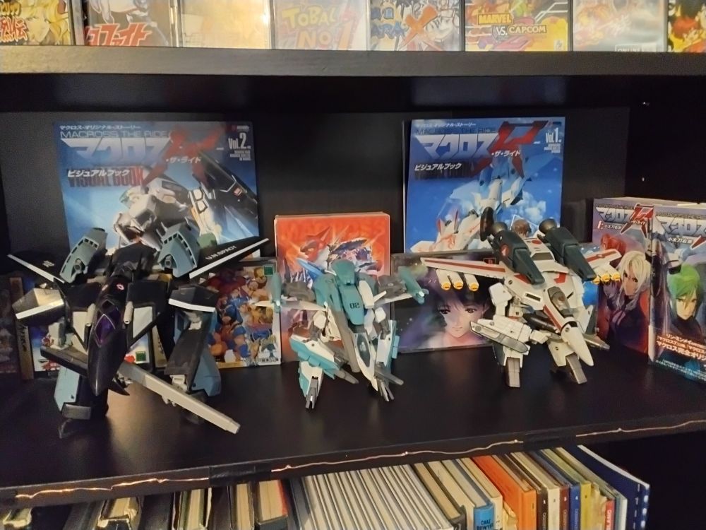 Photo of a shelf mostly dedicated to #Macross.
There’s a VF-17 Black Diamond from Yamato, a VF-2 SS from Bandai and a VF-1 J from Yamato. They’re displayed in Gearwalk or "Ostrich" mode with the rifle in hand and the fastpack equipped.
Behind them are the 2 books and the 2 modeling revues or "Visual Books" for #MacrossTheRide, and the videogames "Macross Do You Remember Love" for PS1 and the collector edition of "Macross 30" for PS3.