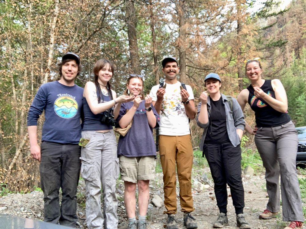 Six happy mycologists standing in a line showing off their favorite finds of the day. In the background is a dry, mostly brown, recently burned forest.