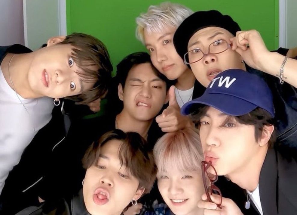 A playful group photo of all seven BTS members shows them making silly faces and posing close together. Jungkook tilts his head with wide eyes, Taehyung winks while pointing towards the camera, J-Hope smiles with an arm around V, RM adjusts his glasses with a surprised expression, Jin puckers his lips while holding sunglasses, Jimin sticks his tongue out, and SUGA smiles brightly. The background is a simple green screen, giving a fun and spontaneous vibe to the photo.