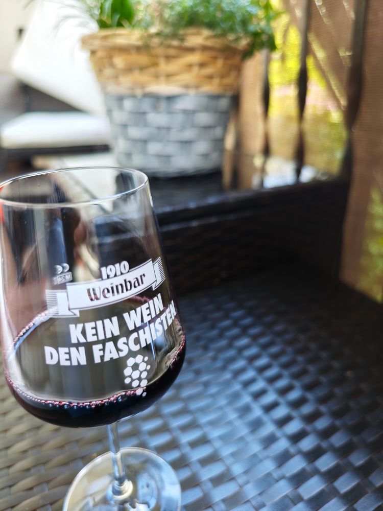 Ein Glas Rotwein auf einem Hocker. Das Glas trägt die Aufschrift, kein Wein den Faschisten.