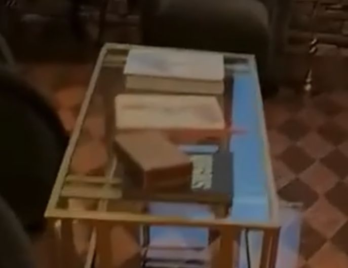 A glass coffee table with a wooden box (perhaps for cigars), 3 books on top and 2 books below. One has a title but is not legible and is partially obscured by the wooden box atop it. The two books on top have cover designs that look like starling mumurations though one might be a large zayin from the Hebrew alefbet.
