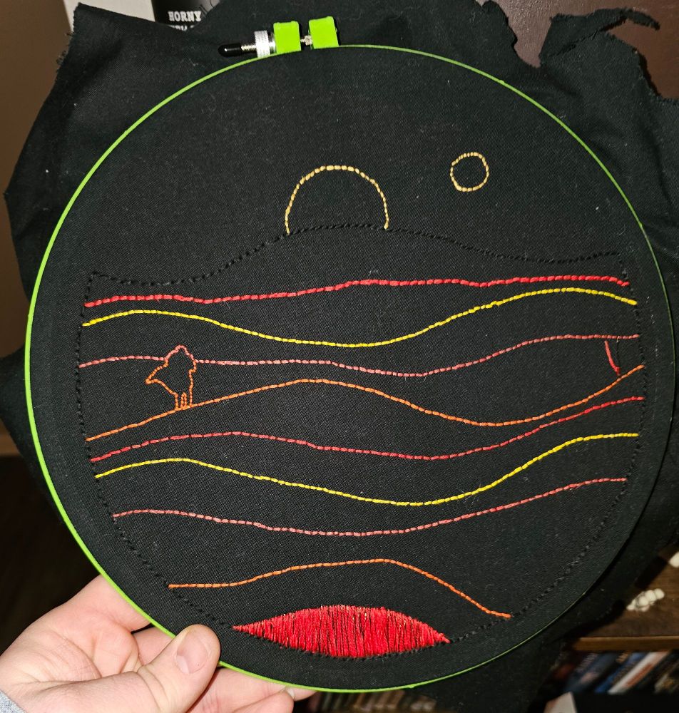 WIP embroidery project of the cover art for Dune. A circle of black fabric showing rolling hills of sand with line of red maroon orange and yellow with a lone wanderer walking into the double moonset. The bottom section is finished in a satin stitch of red with dark copper thread mixed in for the Spice. 