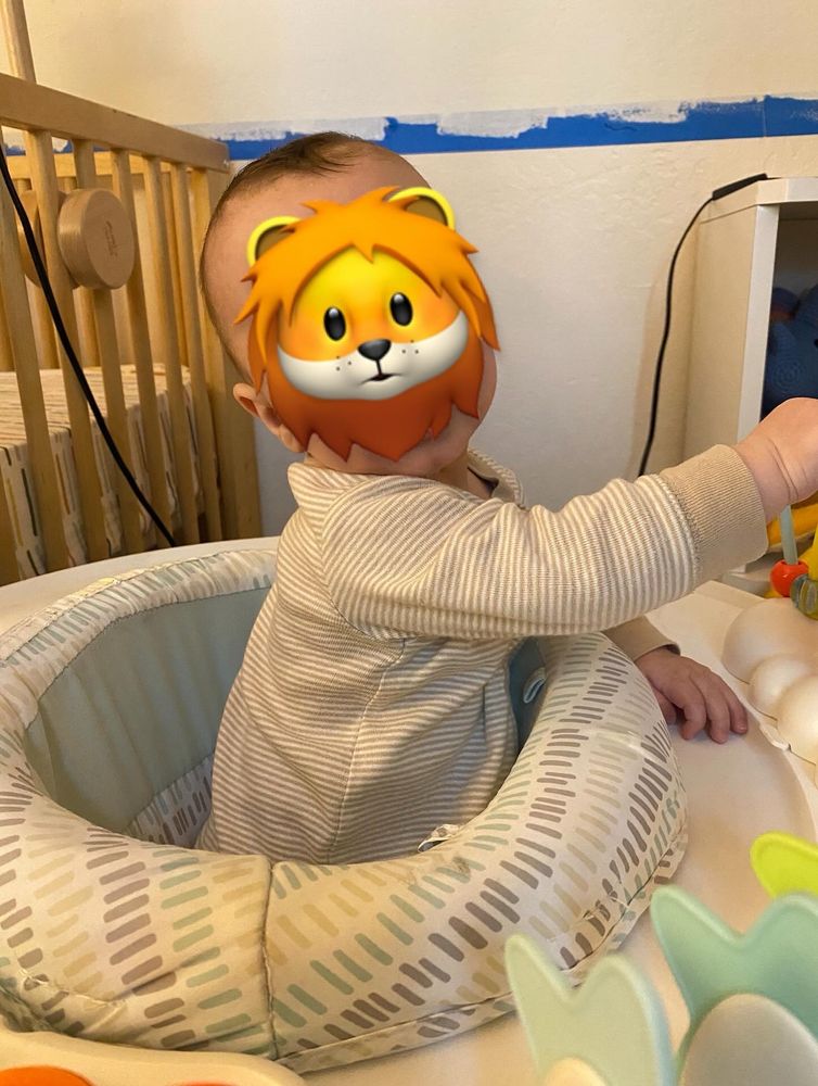 An infant in a baby seat looks towards the camera but their face is obscured by a lion emoji 