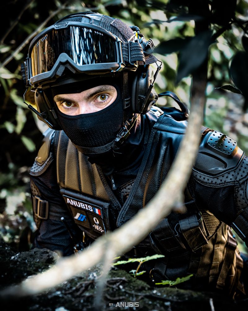 Dressed in black tactical gear, Anubis crouches low in the foliage. His eyes, sharp and visible beneath the helmet and mask, lock onto the target.