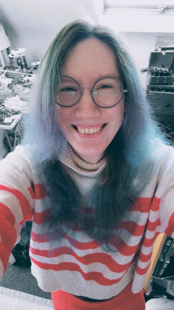 Photo of the author in festive getup, blue hair, red and white striped jumper and red wooly skirt. She is wearing round glasses and smiling. 