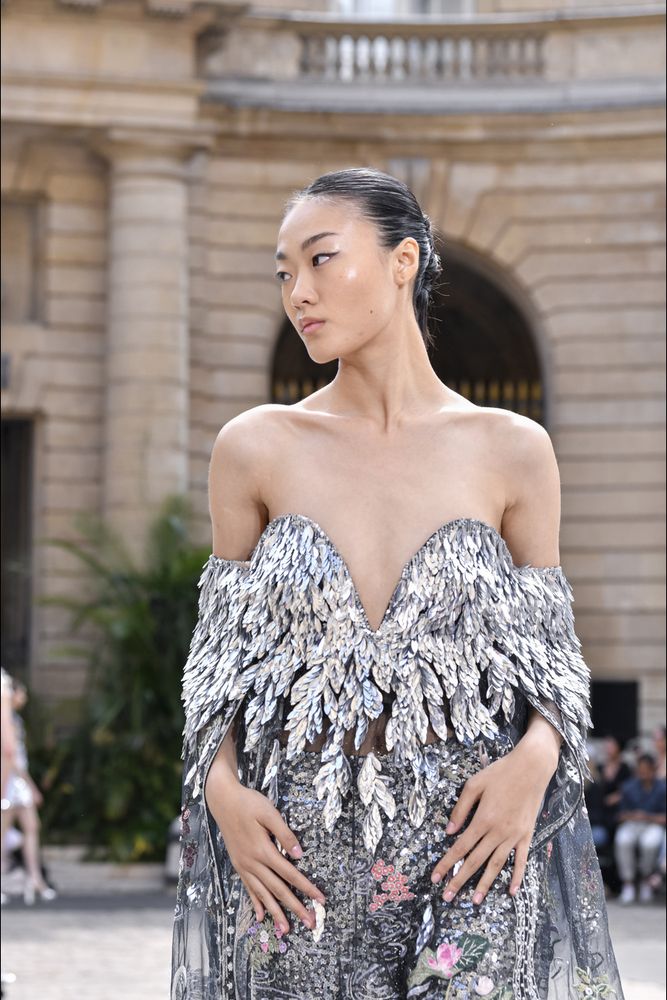 Rahul Mishra strapless top (or bodice) decorated with silver sequins in the shape of leaves or feathers. The sleeves wrap around the bicep/upper arm and extend into a tulle-like cape down the back, also with sequin detailing. The bottoms also use silver sequins, along with pink and green to make small flowers. The model's hair is slicked back into a bun, and she has a fairly minimal makeup look that uses black winged eyeliner, a pinky nude lip, and highlighted cheekbones.