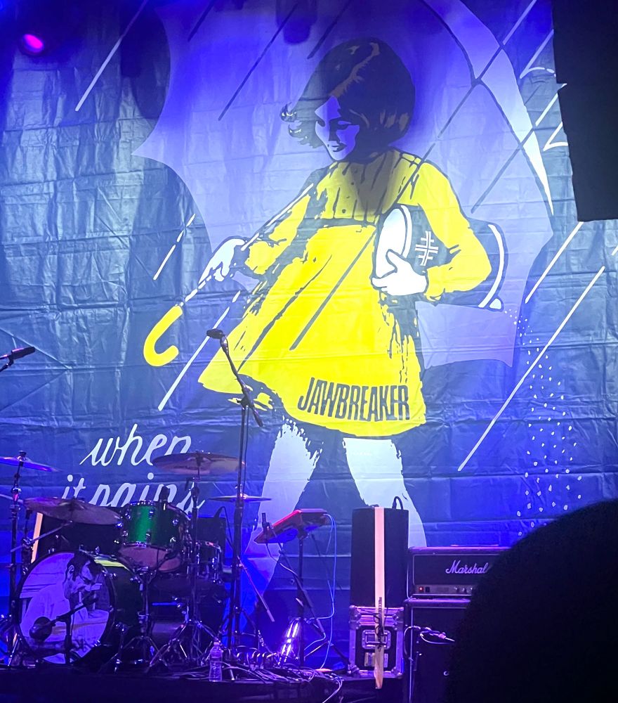 Picture of the stage at the house of blues with Jawbreaker’s instruments set up and being tuned. There’s a big backdrop featuring the Morton’s salt girl (iconic image of little girl wearing a yellow raincoat carrying an umbrella )and it says when it pains it roars instead when it rains it pours. 