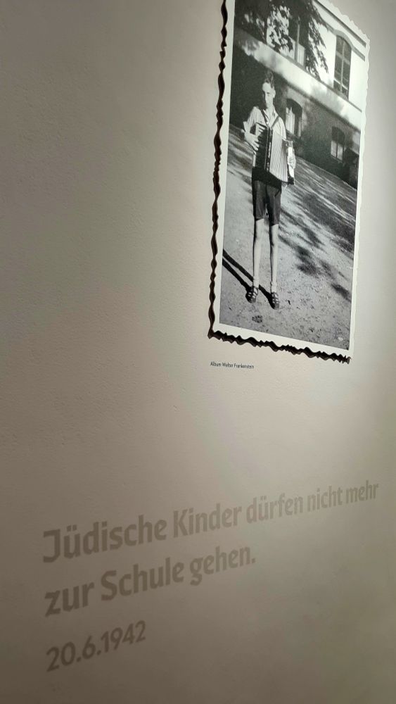 Eine Wand in der Ausstellung. In Hellgrau: Jüdische Kinder dürfen nicht mehr zur Schule gehen. 20.6.1942. Darüber ein Foto von einem Jungen, der Akkordeon spielt. 