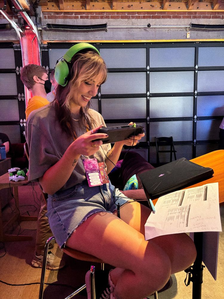 A photo of Ellie sat on a high stool playing on a steam deck and wearing over ear headphones. She is completely engrossed in the game and grinning widely 