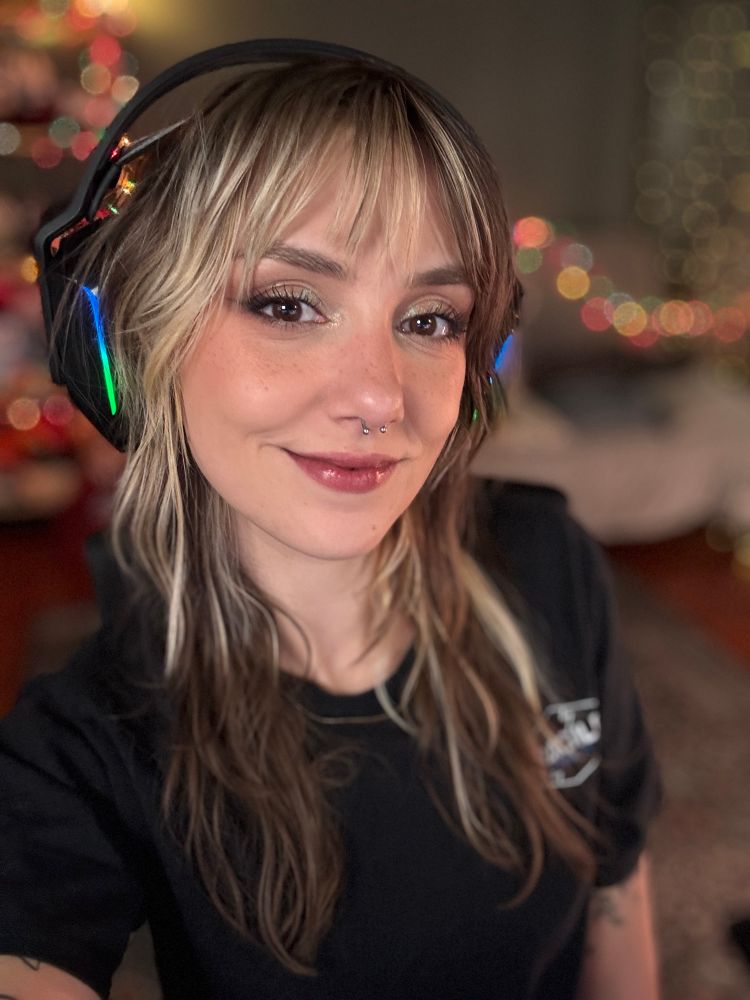 Ellie is smiling in a selfie with her brown and blonde hair loose and wavy. She is wearing an over ear headset and a black t shirt 