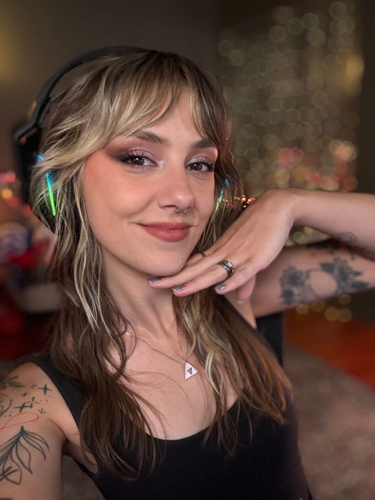 Ellie is smiling in a selfie with her brown and blonde hair loose and wavy, holding her hand gently under her chin. She is wearing an over ear headset and a black tank top and has illustrative linework tattoos on her arms 