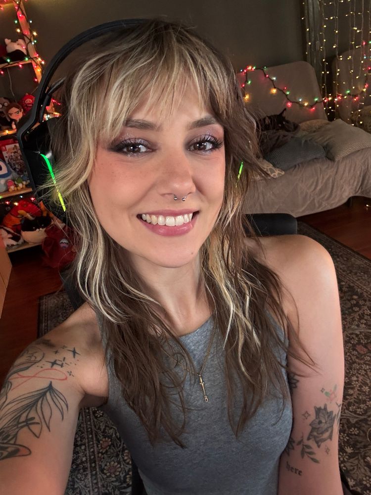 Ellie is smiling in a selfie with her brown and blonde hair loose and wavy. She is wearing an over ear headset and a gray tank top, and has illustrative linework tattoos on her arms 