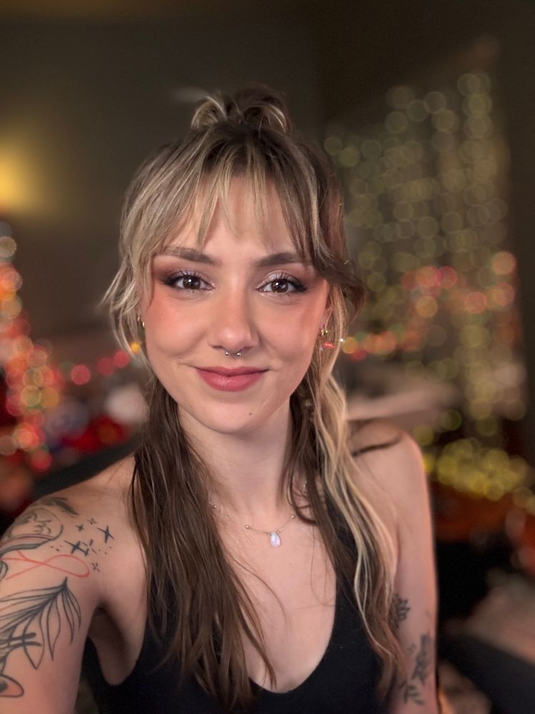 Ellie is smiling in a selfie with her brown and blonde hair tied half up in a bun. She is wearing a black tank top and has illustrative linework tattoos on her arms 
