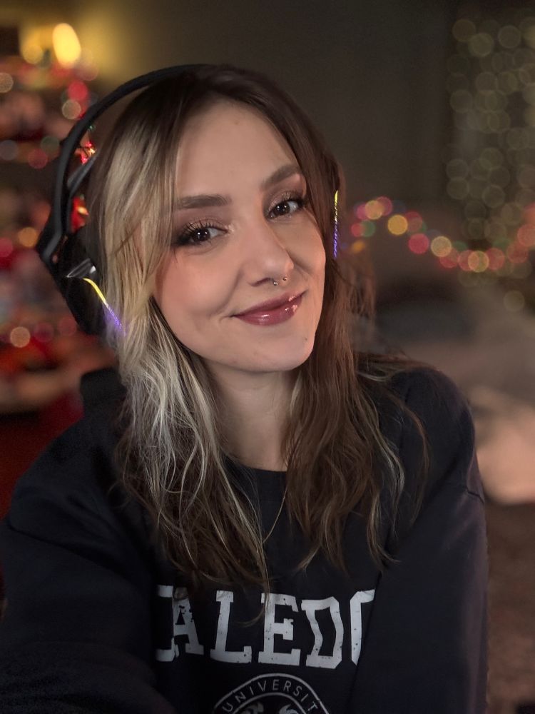 Ellie is smiling in a selfie with her brown and blonde hair loose and wavy. She is wearing an over ear headset and a navy sweater