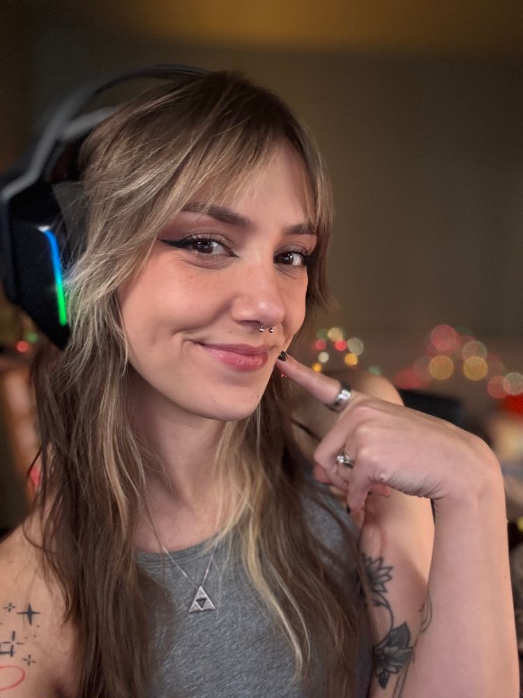 Ellie is smiling cheekily and poking her cheek with a finger in a selfie 