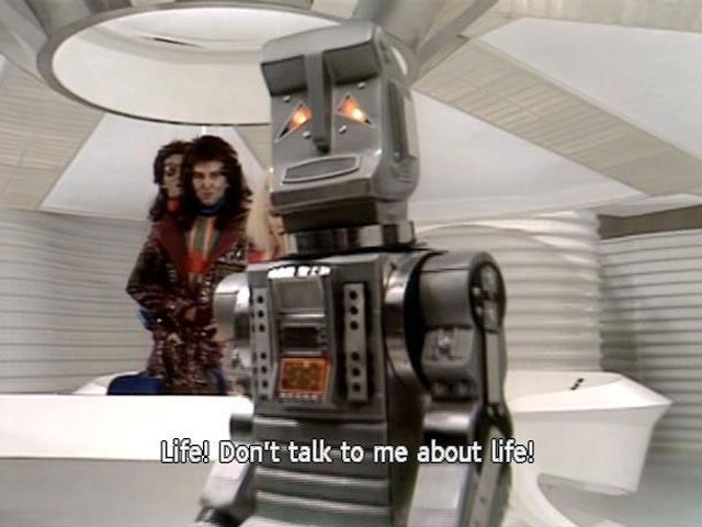 In the clean white interior of the Heart of Gold spaceship, Marvin the paranoid android from hitchhiker’s guide to the galaxy, saying “Life. Don’t talk to me about life!” Zaphod Beeblebrox stands behind with a disapproving look on his faces. 