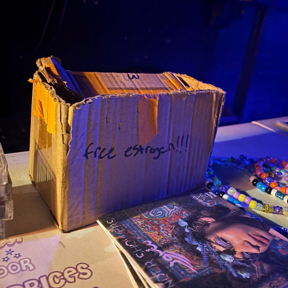 a merch table with a rectangular cardboard box with “free estrogen!!!” written on it in black sharpie marker 