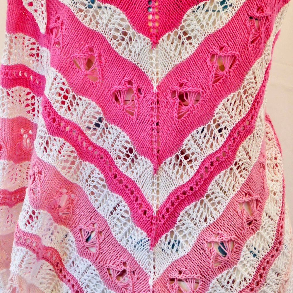 Close-up photo of a knitted shawl with some intricate lace. Stripes of progressively fading shades of pink are interspersed with stripes of white open lace sections. The main lace pattern in the coloured sections are butterfly motifs. The white sections are an airy Feather & Fan lace. 