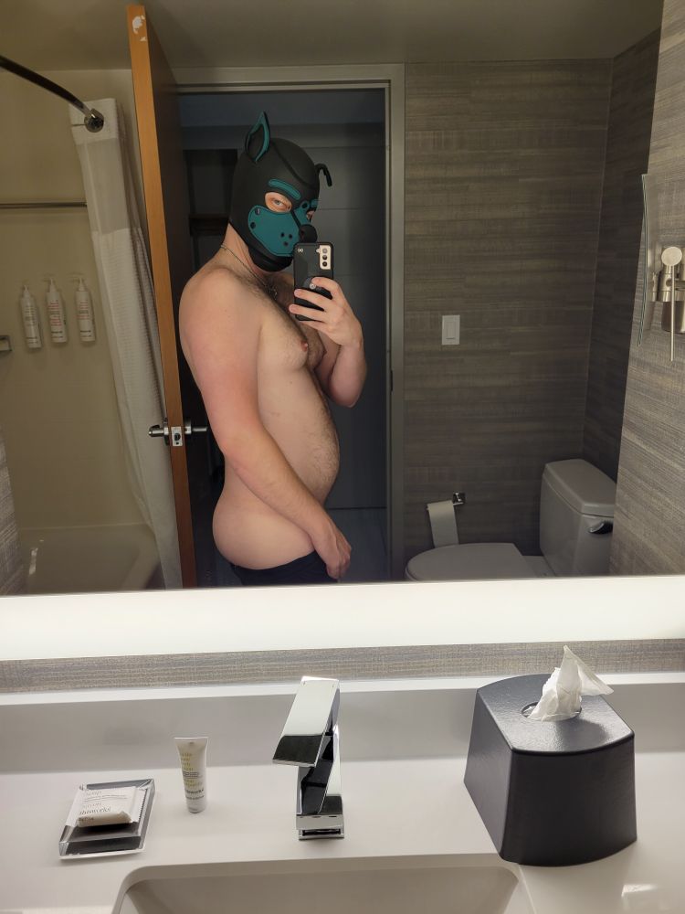 A shirtless person in a green pup hood, taking a selfie in a hotel bathroom mirror. Their ass is out!