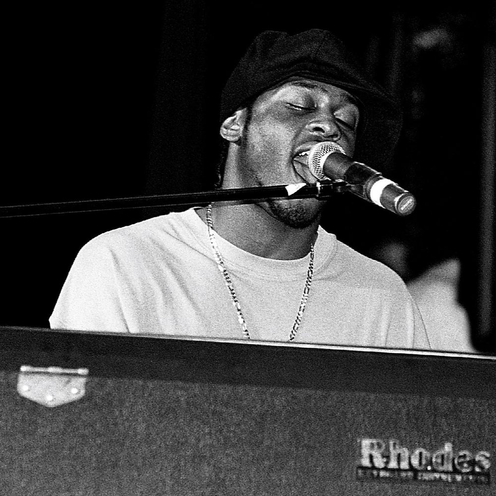 Black-and-white image of D’angelo sitting behind his rhodes, singing into a microphone on a stand which has been swiveled in front of his face as he plays.