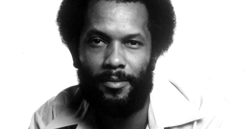 A black and white photo of Roy Ayers, wearing a wide-collared shirt. He stares into the camera confidently, head-on, a slight smile on his lips. It is the face of a man who knows he’s the baddest.