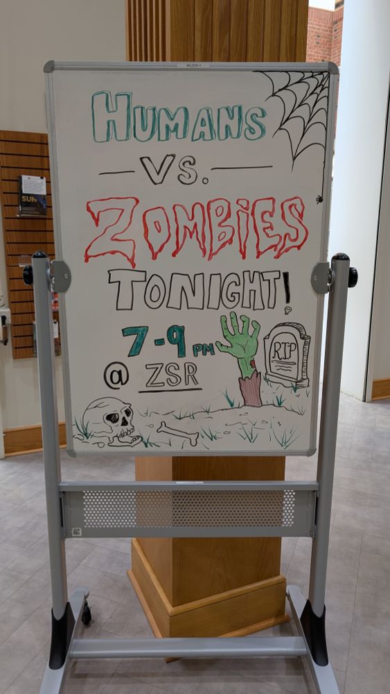 The following test is written on a whiteboard:

Humans vs. Zombies
Tonight!
7-9 p.m.
@ZSR

A spider web, a skull, and a hand rising from a grave marked "RIP" are around the text.