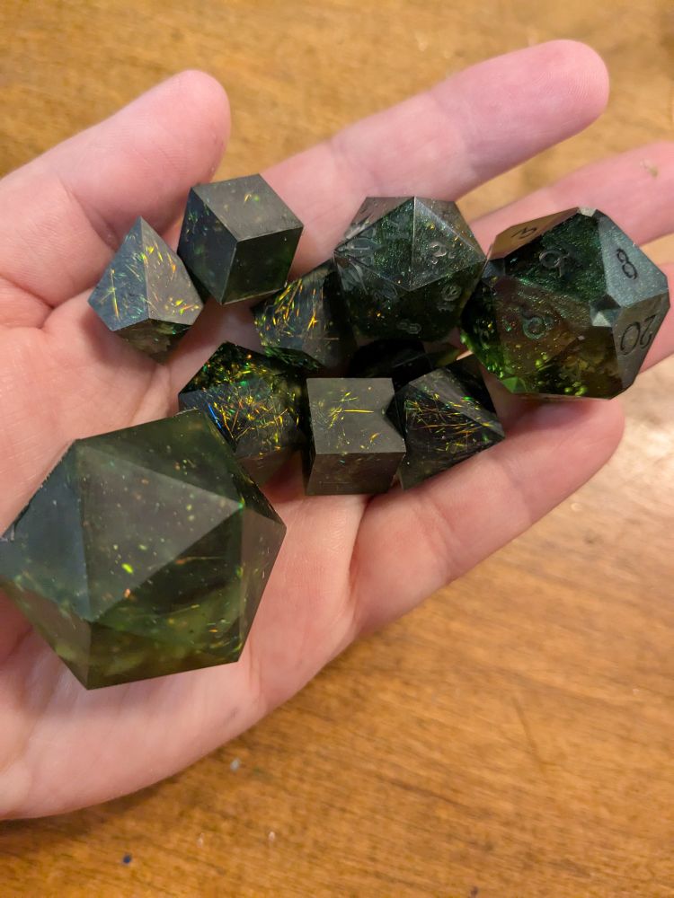 A set of blanks for resin dice made with green resin and fibers that flash yellow, green and orange. There are seven polyhedral dice and a large d20. These blanks do not have numbers yet, but will be cast again in numbered molds. Two d20s were poured using leftover resin, one features inverted corners and the other has flower motifs on some of the faces.